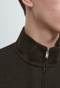 Ciemnobrązowa kurtka z polaru z wysokim kołnierzem, z frontowym zamkiem błyskawicznym i fakturowaną tkaniną. Zbliżenie na kołnierz i okolice szyi.