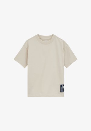 Beżowy t-shirt z krótkim rękawem i okrągłym dekoltem, z małą granatową naszywką blisko dolnego brzegu po prawej stronie z przodu.