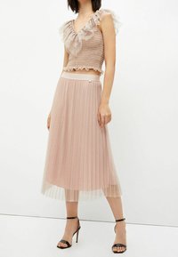 Mujer luciendo un top corto con volantes en color rosa empolvado y una falda midi plisada brillante, combinada con sandalias de tacón alto negras.