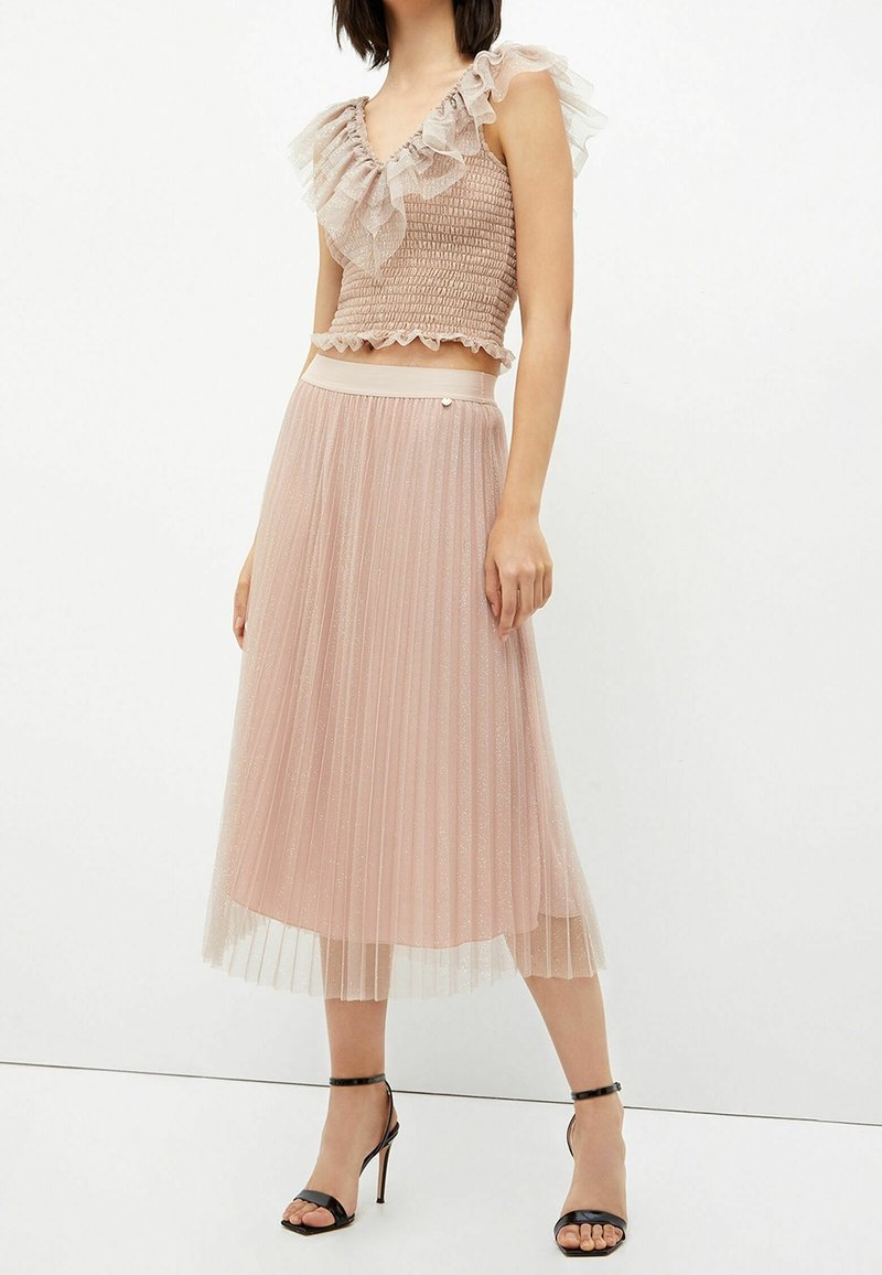 Mujer luciendo un top corto con volantes en color rosa empolvado y una falda midi plisada brillante, combinada con sandalias de tacón alto negras.