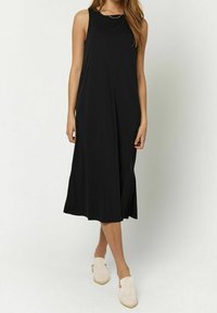 Robe sans manches noire en tissu doux, présentant un col rond, un design fluide et des fentes latérales, associée à des chaussures de couleur claire.