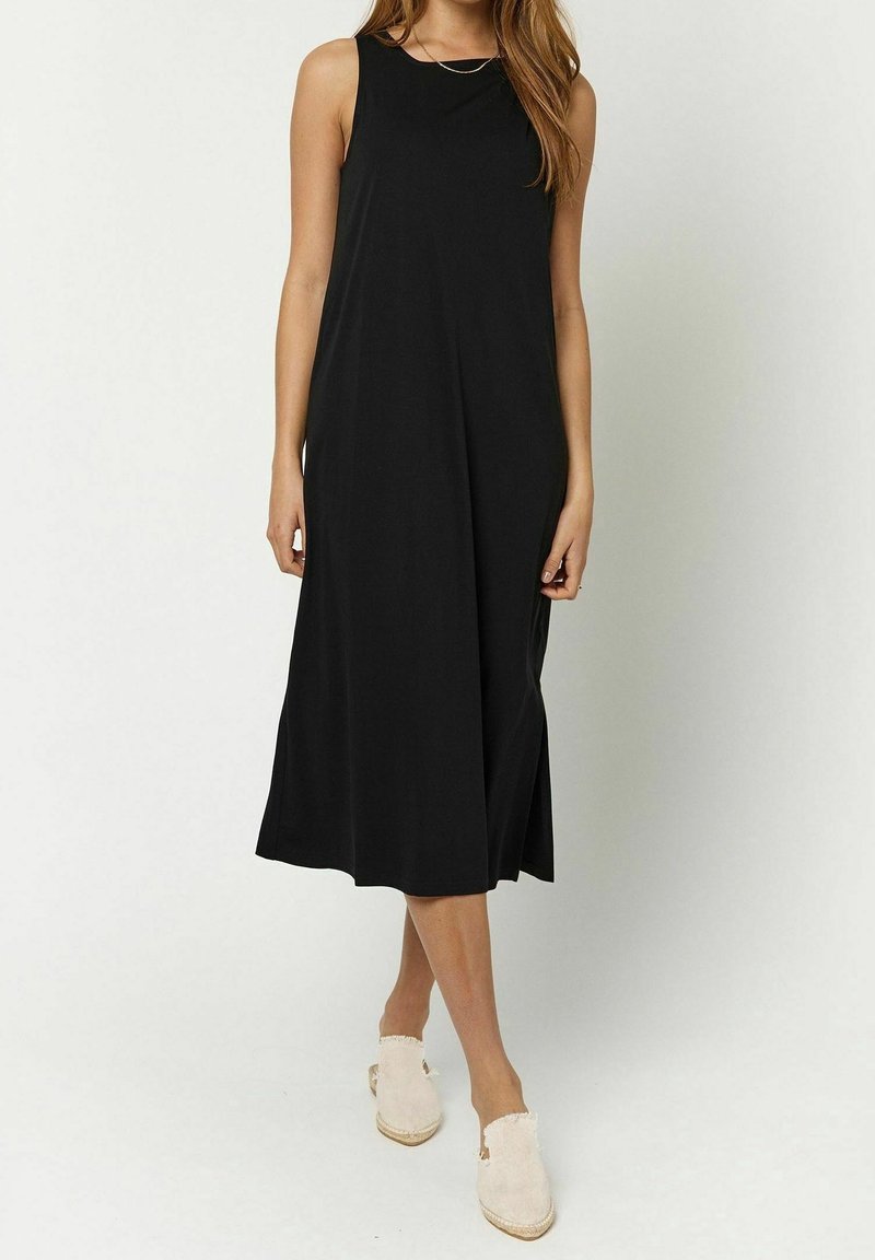 Robe sans manches noire en tissu doux, présentant un col rond, un design fluide et des fentes latérales, associée à des chaussures de couleur claire.