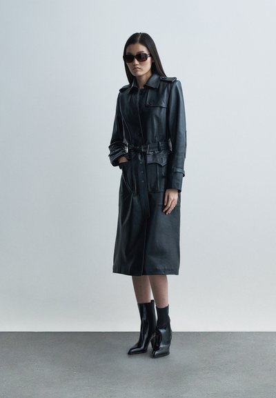 Juodas odinis trapecinis paltas su diržu ties liemeniu, priekiniais mygtukais ir dviem šoninėmis kišenėmis. Derinamas su prigludusiais juodais auliniais batais.