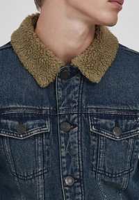 Džinsa jaka ar bēša, pūkainu apkakli, tumši zilu nokrāsu, divām krūšu kabatām ar pogām un klasisku pogu aizdari priekšpusē.