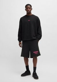 Felpa nera con logo rosa, abbinata a pantaloni corti neri con testo rosa. Caratterizzata da una vestibilità rilassata e un materiale morbido; le scarpe sono nere.