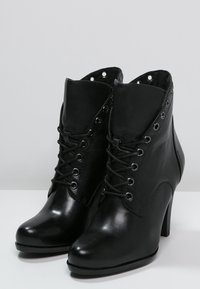 Botines negros hasta el tobillo con un acabado de cuero brillante, punta redonda, tacón alto y ojales decorativos a lo largo del cordón. Cuenta con un caña acolchada.