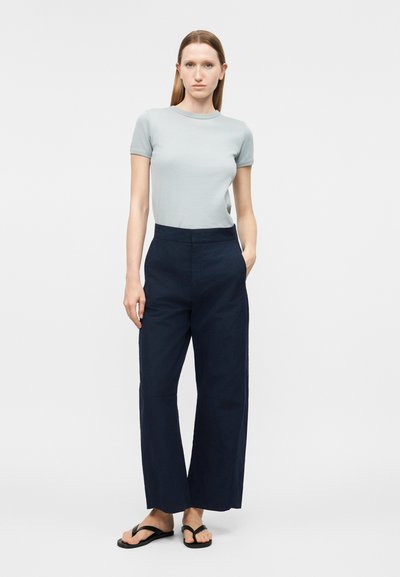 Γυναίκα που στέκεται, φοράει ανοιχτό γκρι κοντομάνικη μπλούζα, μπλε σκούρο φαρδιά παντελόνια και μαύρα σανδάλια τύπου φλιτ-φλοπ μπροστά σε μονόχρωμο φόντο.