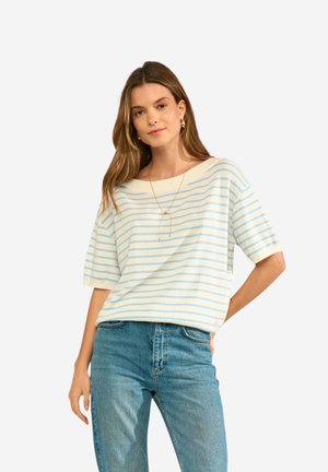 Młoda kobieta ubrana w biały i jasnoniebieski pasiasty krótki top z krótkim rękawem oraz wysokie niebieskie dżinsy, stojąca z jedną ręką w kieszeni.