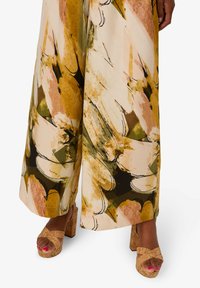 Kalhoty s širokými nohavicemi mají živý abstraktní potisk v odstínech žluté, růžové a zelené, doplněné krátkými sandály v béžové barvě s kříženými pásky.