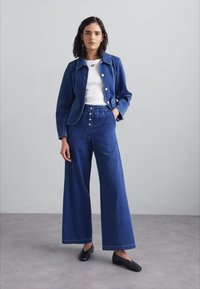 Kurtka dżinsowa z zapięciem na guziki, w połączeniu z dżinsami o szerokich nogawkach i wysokim stanie. Oba elementy w ciemnoniebieskim dżinsie. Pod spód biała bluzka i czarne baleriny.