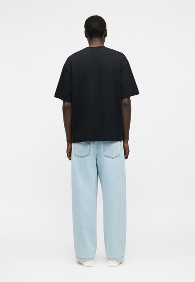 Άτομο φαίνεται από πίσω φοράει μαύρο κοντομάνικο πουκάμισο, ανοιχτό μπλε τζιν με φαρδιά πόδια και λευκά αθλητικά παπούτσια πάνω σε απλό φόντο.