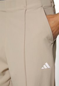 Beige Sporthose aus weichem Stoff, mit Seitentasche und weißem Adidas-Logo-Akzent am unteren Bein. Glatte Textur.