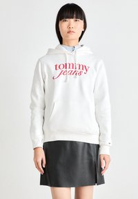 Vit hoodie med "tommy jeans" i röd skrift, med en framficka och dragsko vid huva. Parat med en svart läderkjol.