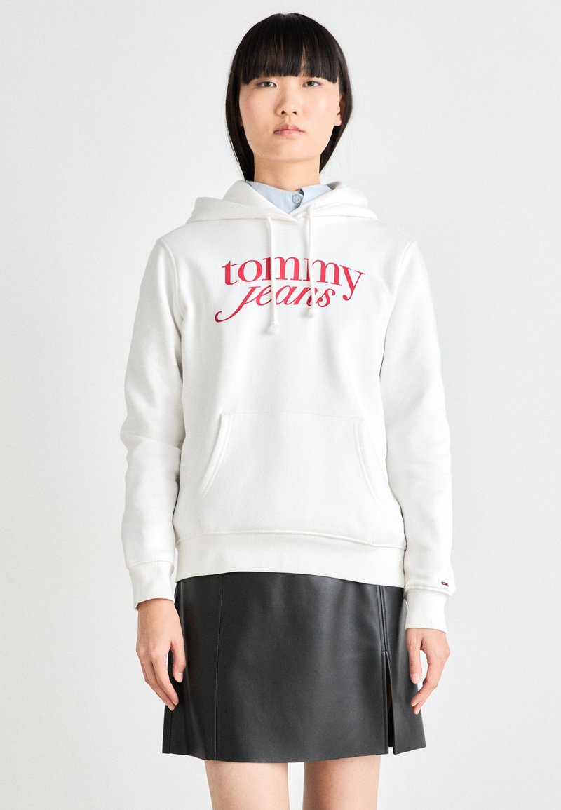 Vit hoodie med "tommy jeans" i röd skrift, med en framficka och dragsko vid huva. Parat med en svart läderkjol.