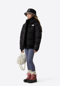 Svart dunjakke, lyseblå leggings, røde støvler med hvit pelskant og en kremfarget ryggsekk. Har en svart bøttehatt med grå fôr.