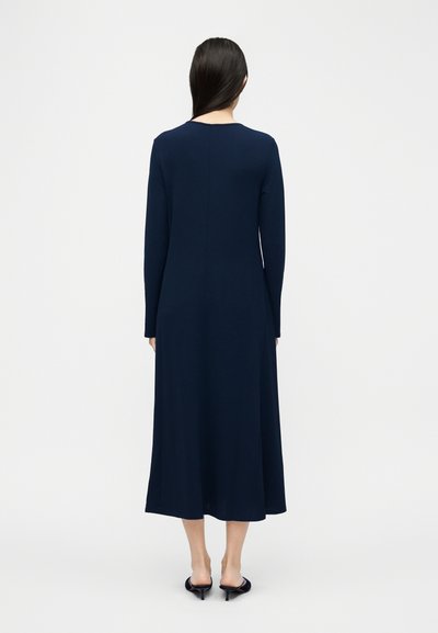 Γυναίκα με μακριά σκούρα μαλλιά, φοράει μακρυμάνικο φόρεμα μέσης μήκους σε ναυτικό μπλε χρώμα και μαύρα ψηλοτάκουνα mules, με την πλάτη στραμμένη σε λευκό φόντο.