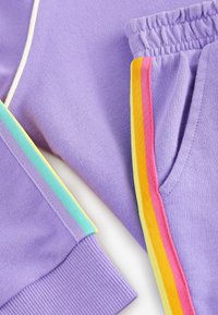 Lavendelfarbene Sweatpants mit elastischem Bund, ausgestattet mit Streifen in Türkis, Gelb, Rosa und Orange an den Seiten. Weicher Baumwollstoff.