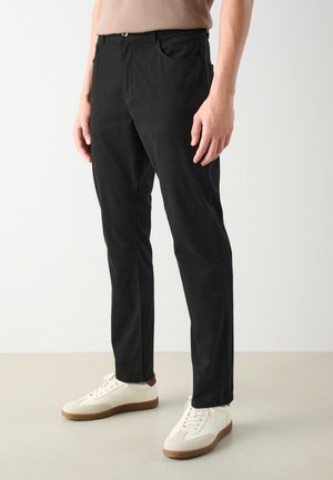 Osoba nosząca wąskie czarne spodnie i białe trampki, stojąca na jasnej podłodze na tle gładkiej białej ściany.