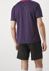 Lila idrottströja med rosa krage, korta ärmar och textilstruktur, kombinerad med svarta shorts, visad bakifrån.