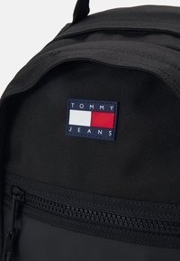 Mochila de tela negra con un parche azul marino prominente que tiene rectángulos blancos y rojos, etiquetado como "TOMMY JEANS", y un compartimento con cremallera.