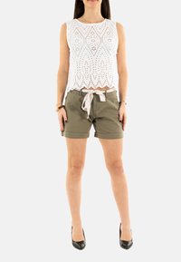 Top blanco sin mangas con patrones recortados; pantalones cortos verde oliva con un cinturón de tela; modelo con tacones altos negros; en posición de pie.