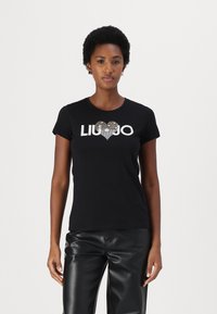 Mustat t-paidat, joissa on lyhyet hihat, ja joissa on hopeinen "LIU JO" -logo ja sydämen muotoilu, jotka on koristeltu strasseilla. Yhdistetty mustiin nahkahousuihin.