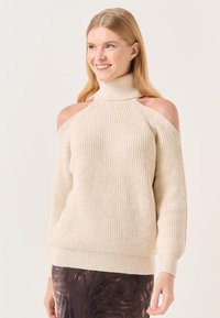 Pull en tricot beige avec un col montant, épaules dénudées et texture côtelée. Manches longues et coupe décontractée, associé à une jupe à motifs.