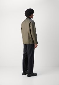 Chaqueta ligera verde olivo con cuello, bolsillos laterales y detalles en botones, llevada sobre pantalones oscuros y zapatos negros. Vista trasera.