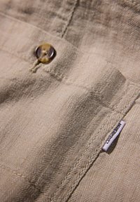 Tela en espiga beige con una superficie texturizada, que presenta un botón marrón y una pequeña etiqueta con "LINDENBERGH" en el borde.