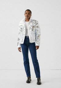 Adult wearing white sequin puffer jacket, white shirt, blue jeans, and black boots standing against a plain background.