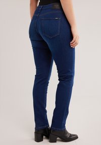 Isik kandmas tumesiniseid kitsaid teksapükse ja musti kontsaga pahkluu-saapaid, seisab ühe käega kehapoolel.