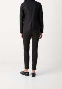 Chaqueta negra a medida con un corte ajustado, textura suave y puños con botones, combinada con pantalones oscuros de corte slim y zapatos negros sin cordones.