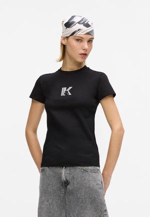 Femme portant un t-shirt noir moulant avec un logo argenté, un foulard imprimé sur la tête et un jean en denim gris taille haute, debout devant un fond blanc uni.