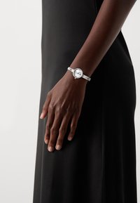 Silver wristwatch with a round white dial, minimalist face, and textured metal band. Adjustable clasp and subtle decorative accents.