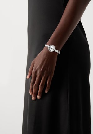 Silver wristwatch with a round white dial, minimalist face, and textured metal band. Adjustable clasp and subtle decorative accents.
