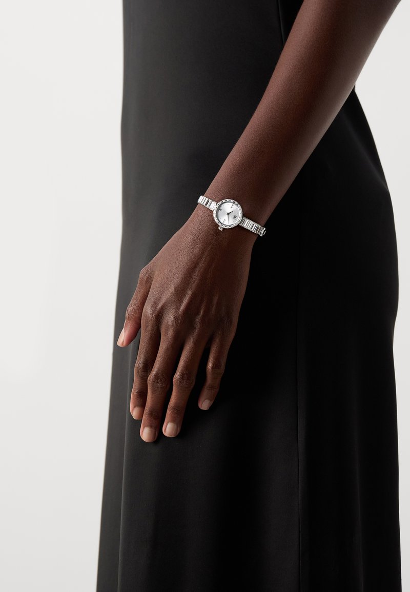 Silver wristwatch with a round white dial, minimalist face, and textured metal band. Adjustable clasp and subtle decorative accents.