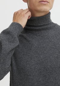 Pull à col roulé gris foncé en tissu tricoté texturé. Il présente un col et des poignets côtelés, avec une coupe ajustée.
