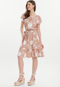 Vestido blanco con un patrón de paisley en rosa y dorado, mangas cortas abullonadas, cintura con cordón y un dobladillo de volantes en capas. Llevado con sandalias de cuña beige.