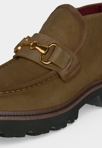 Mocassin en daim marron avec détail en métal doré en forme de mors de cheval et semelle en caoutchouc noire épaisse, montrant le dessus et le côté avant.