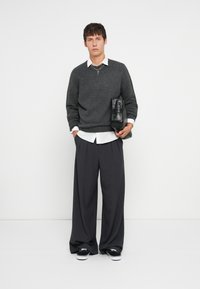 Grijze trui met een V-hals, een witte blouse eronder, losse grijze broeken en zwarte sneakers. Houdt een zwarte tas vast met hardware details.