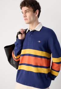 Marineblå langermet polo med kremfarget krage, med horisontale striper i gul, oransje og blå. Designet inkluderer en liten logo.
