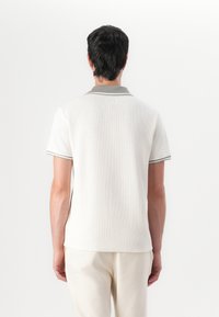 Kurzärmliges Polohemd in Creme mit strukturiertem Stoff, grauem Kragen und kontrastierenden Akzenten entlang der Ärmel. Kombiniert mit hellen Hosen.