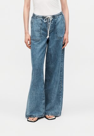 Nő magas derekú, világoskék, bőszárú farmert visel elülső foltzsebekkel és fehér zsinórral, fehér felsővel és fekete, négyzetorrú szandállal párosítva.