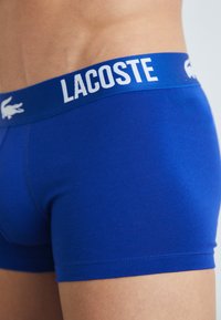 Zili bokseri, izgatavoti no gludas kokvilnas, ar baltu elastīgu jostu, kas rotāta ar "LACOSTE" logotipu un krokodila emblēmu.