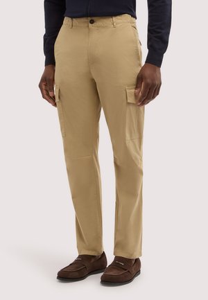 Mężczyzna w beżowych spodniach typu cargo i brązowych mokasynach z zamszu, stojący z rękami swobodnie opuszczonymi wzdłuż ciała na neutralnym tle.