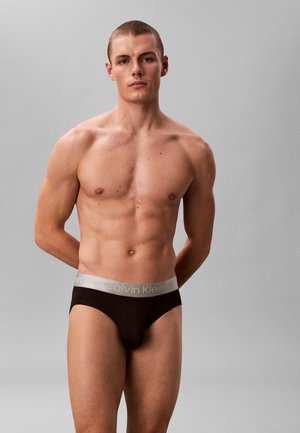 Jeune homme debout, mains derrière le dos, portant un slip noir Calvin Klein sur un fond gris uni.