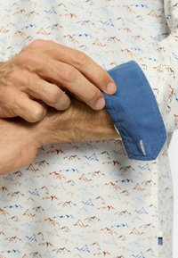 Weißes Hemd mit einem Bergmuster in Blau-, Braun- und Rottönen. Die blaue Manschette besteht aus geripptem Material und enthält ein Etikett. 