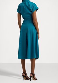 Tealfärgad bomullsjacka med korta, upprullade ärmar, en figursydd överdel och en utsvängd kjol. Matchad med svarta, remmar högklackade skor.