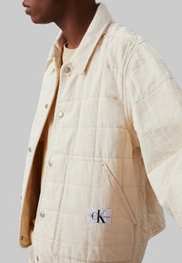 Giacca trapuntata beige con colletto, bottoni a pressione e tasche frontali. Presenta un'etichetta con il marchio "Calvin Klein Jeans" sul lato.