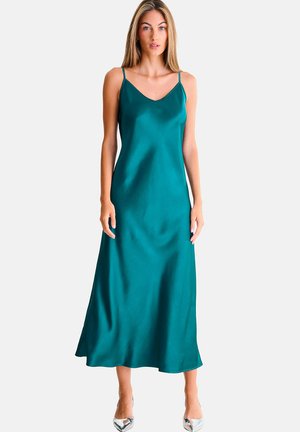 Femme portant une robe nuisette en satin coloris bleu sarcelle avec des bretelles fines et des talons argentés à bout pointu, debout devant un fond clair uni.